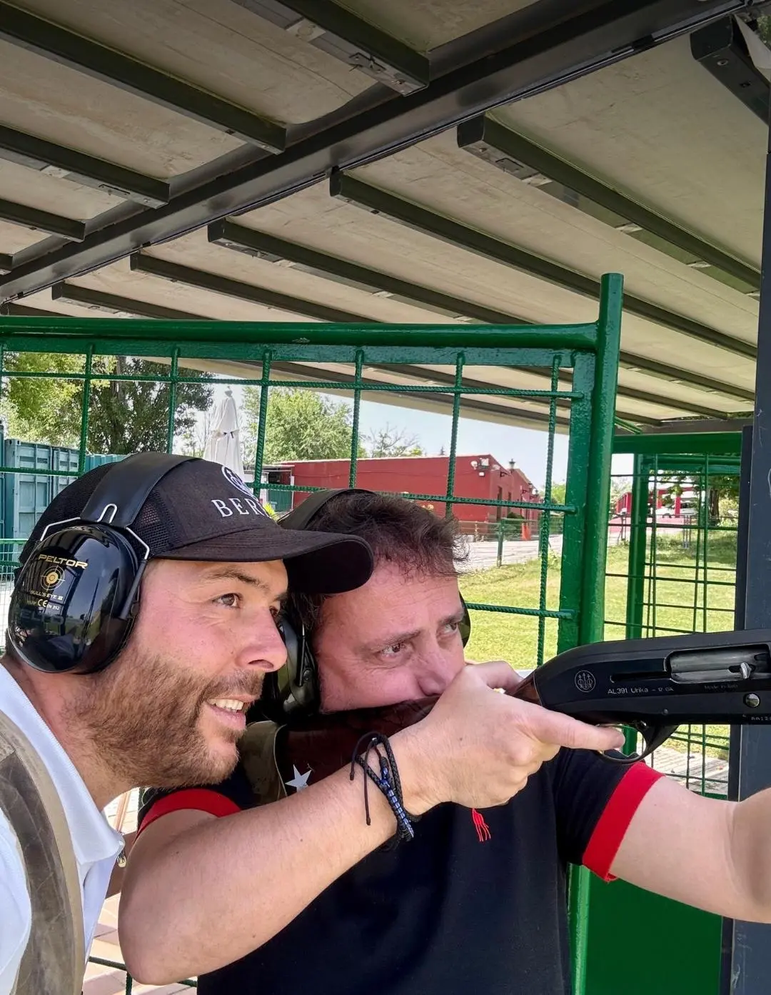 Gonzalo Gómez Arrimadas dando indicaciones técnicas a un alumno durante una clase de tiro deportivo en campo oficial, ambos con protección auditiva.